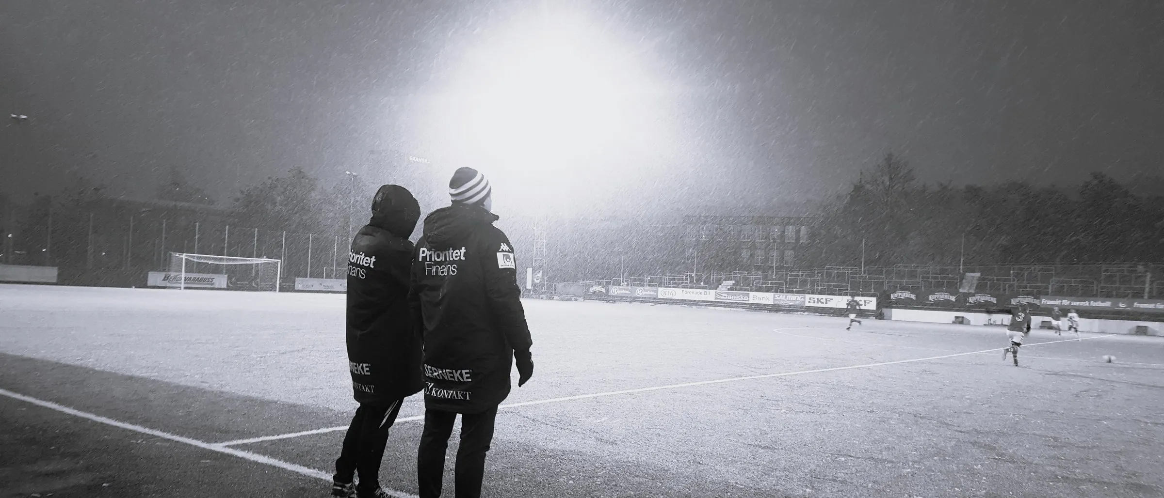Två tränare med ryggen mot kameran vid sidlinjen under en match i snöfall, i strålkastarljus, med spelare ute på planen.